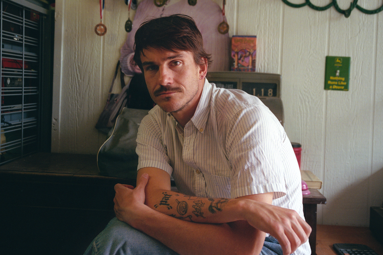 Musician Medium Build (Nick Carpenter) wearing a striped shirt and showing arm tattoos while sitting indoors for a casual press portrait photo.
